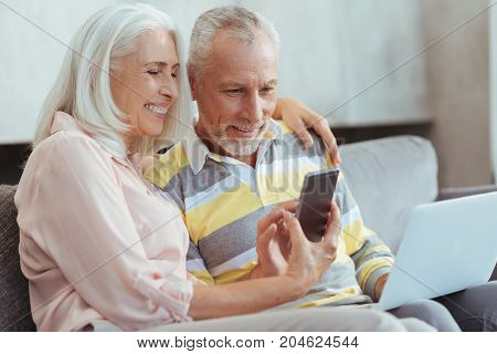 In pace with modern world. Waist up of a cheerful aged couple sitting on the couch and using modern devices while resting at home