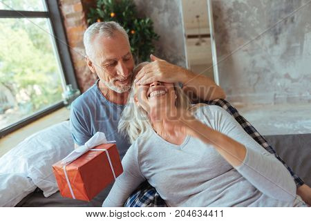 Close your eyes. Pleasant aged man closing eyes of his beautiful wife and holding a present while going to congratulate her