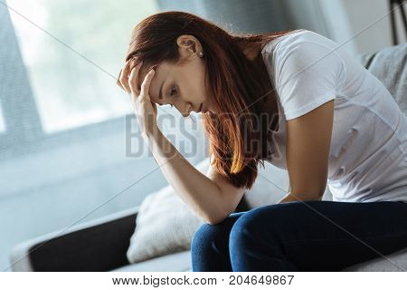 Tough problem. Cheerless upset unhappy woman sitting on the sofa and holding her forehead while looking for a solution of her problem