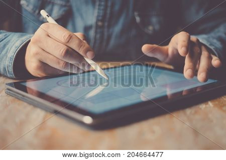 Man working contemporary electronic tablet while sitting at the wooden table at office.Men using digital stylus for pointing device screen.Blurred background. Horizontal
