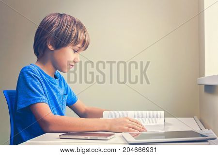 Focused boy reading a book. Tablet computer and mobile phone near to him at the table at home