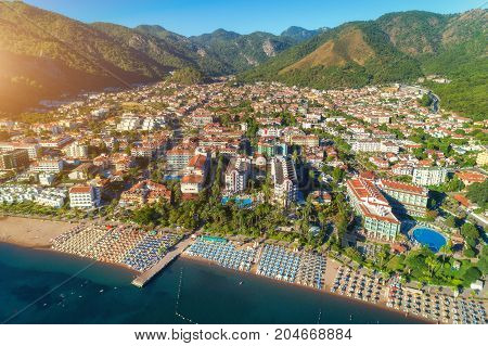 Aerial view of beautiful city, sandy beach with chaise-lounges, mountains, green trees, hotels, buildings, pool, at sunset in Icmeler, Turkey. Summer cityscape Top view from drone. Architecture