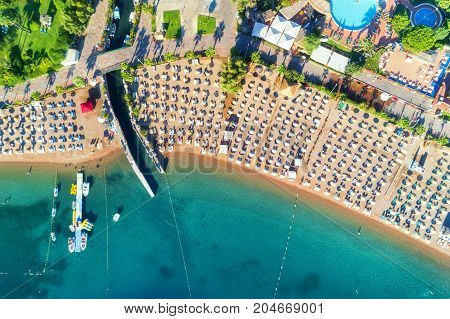 Aerial view of transparent turquoise sea, beautiful sandy beach with colorful chaise-lounges, boats, green trees, pool, hotels, buildings at sunrise in Icmeler, Turkey. Summer landscape. Top view.