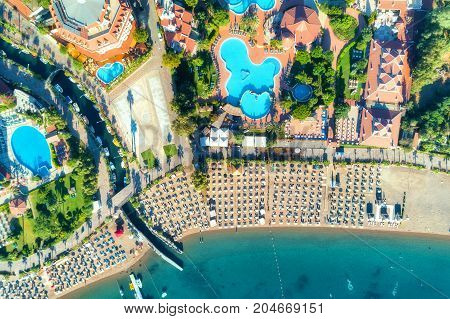 Aerial view of transparent turquoise sea, beautiful sandy beach with colorful chaise-lounges, boats, green trees, pool, hotels, buildings at sunrise in Icmeler, Turkey. Summer landscape. Top view.