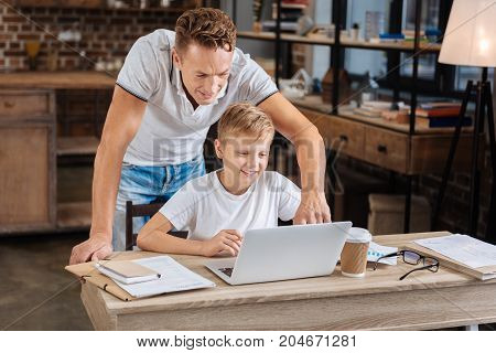 Being helpful. Charming young man standing behind his pre-teen son and pressing a keyboard button on his sons laptop, helping him with laptop settings