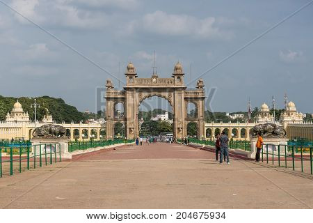 Mysore India - October 27 2013: Brown stone main East Gate to Mysore Palace with entire yellow side buIldings under cloudscape. Visitors add colors.