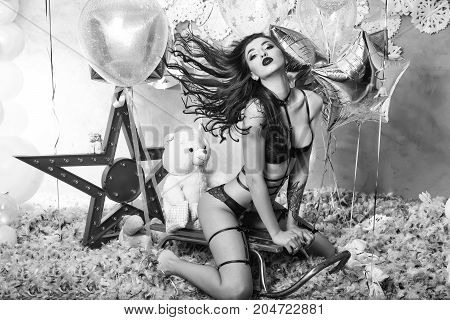 pretty woman or cute sexy girl in erotic lingerie of bra panties has red lips sitting on sledge at teddy bear toy festive golden balloons white snowflakes feathers. party and holiday celebration