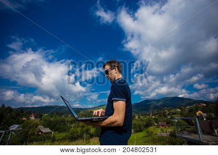 A young man working on a laptop outdoors high in the mountains with a beautiful sky in the background
