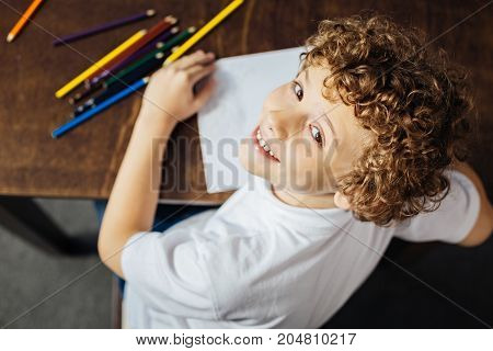 Hello there. Top view on a curly haired boy looking into the camera with a cheerful smile on his face while sitting at a table and drawing at home.