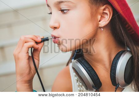 little schoolgirl puts the plug into her mouth. child in a red hat, white blouse and with grey headphones on the background of stairs