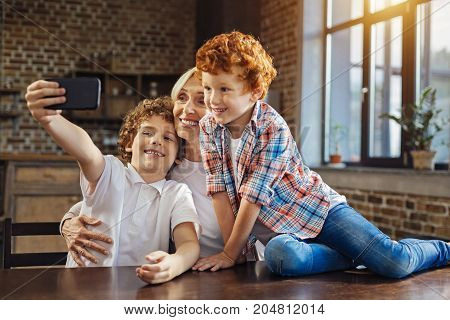 Family memories. Selective focus on a beautiful elderly woman and a chestnut haired boy grinning broadly while looking at a front camera of a phone and taking selfies at home.