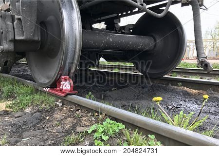 The brake shoe is placed under the wheel of a railway car. A dandelion grows near the rail.