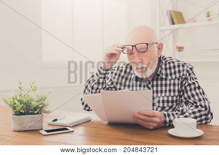 Shocked senior man looking at bills in disbelief, holding his glasses on forehead, copy space