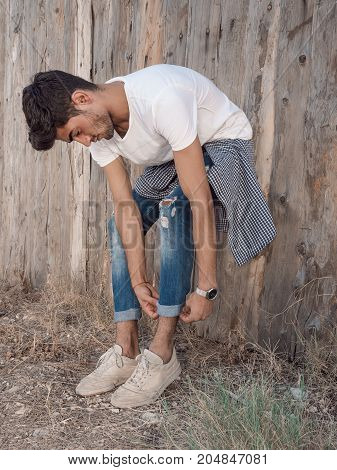 Confident young man in full length standing against wooden wall and correcting clothes. Handsome man with facial hair wearing shirt t-shirt and jeans resting outdoors.