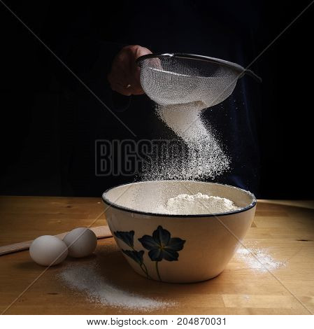 flour falling in a bow from a sieve into a bowl on a wooden kitchen worktop at baking preparation dark background with copy space selected focus