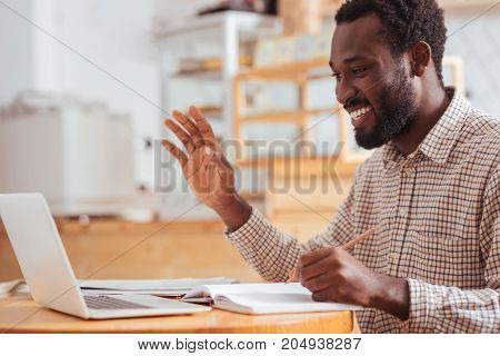 Hello there. Charming cheerful man sitting at the table in the coffee house, making some notes and greeting someone while making a video call