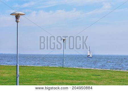 A sailing yacht runs along the shore with a green lawn and slender lanterns. Neringa