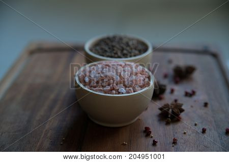 Pink Himalayan salt and coriander seeds in white bowls on wooden surface covered with anise and red peppercorn