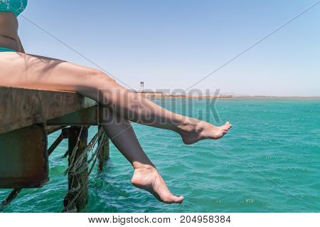 Young Female Legs Hang From The Pier Over The Sea Water