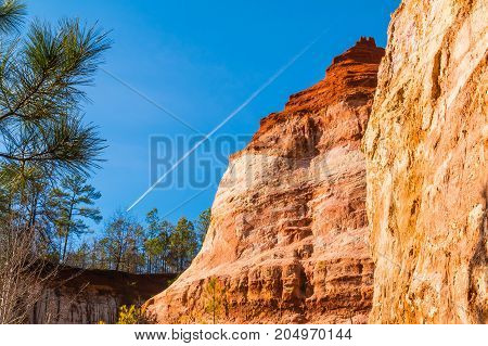 Red loamy wall of Providence Canyon with trees on the background of clear sky with contrail USA