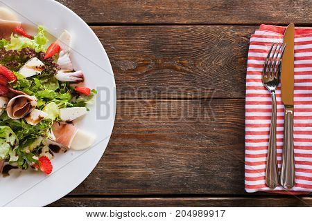 Fresh salad with parma ham serving in restaurant. Lettuce with blue cheese, ruccola and strawberry, gourmet cuisine, top view and free space on dark wooden background