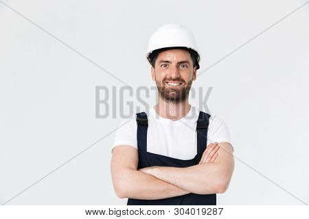 Confident bearded builder man wearing overalls and hardhat standing isolated over white background, arms folded