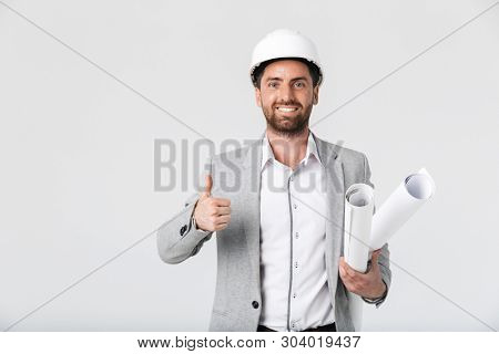 Confident bearded man builder wearing suit and hardhat standing isolated over white background, carrying blueprints, thumbs up