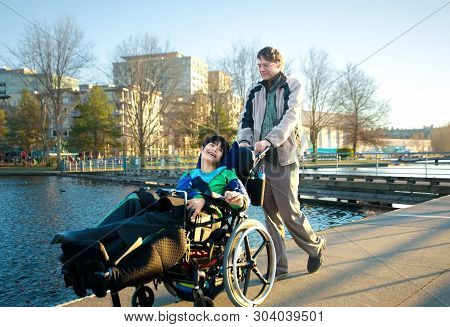 Caucasian Father Walking Disabled Biracial Young Son At Park By Lake Outdoors In Early Spring Or Sum