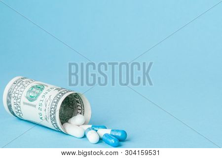 Money Dollar Rolled Up With Pills Flowing Out Isolated On Blue Background, High Costs Of Expensive M