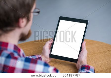 Man Sitting At Wooden Table And Looking At Digital Tablet Computer Device With White Blank Screen. M