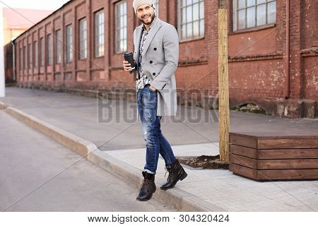 Handsome Young Man In Grey Coat And Hat Crossing The Street With A Cup Of Coffee.