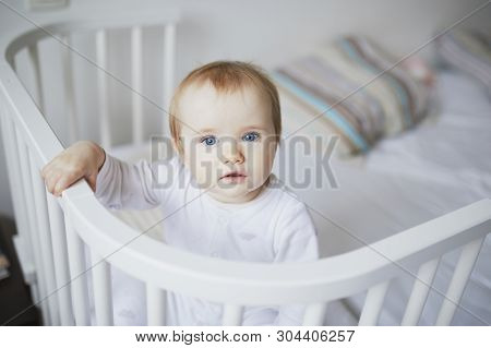 Adorable Baby Girl In Co-sleeper Crib Attached To Parents Bed. Little Kid Sitting In Cot
