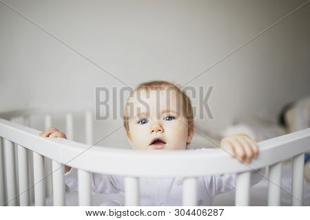 Adorable Baby Girl In Co-sleeper Crib Attached To Parents Bed. Little Kid Sitting In Cot