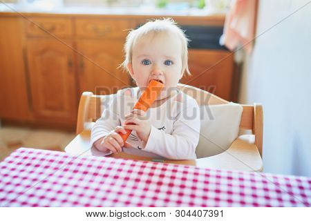 Cute Baby Girl Eating Carrot In The Kitchen. Little Kid Tasting Solids At Home. Baby Led Weaning Con