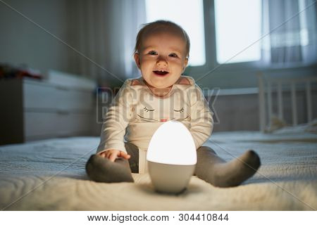 Adorable Baby Girl Playing With Bedside Lamp In Nursery. Happy Kid Sitting On Bed With Nightlight. L