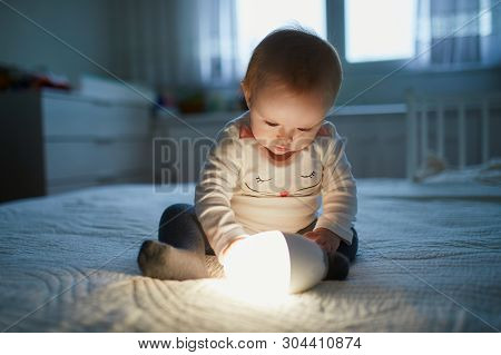 Adorable Baby Girl Playing With Bedside Lamp In Nursery. Happy Kid Sitting On Bed With Nightlight. L
