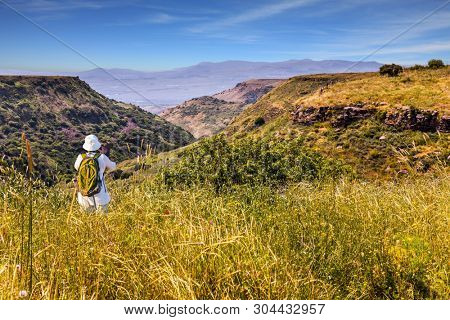 Woman with backpack in white hat photographs picturesque landscape. Jewish Easter in Israel.  Spring blooming Golan Heights - a mountain plateau of volcanic origin. The concept of active and photo 