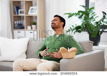 consumption and people concept - full and pleased indian man eating takeaway food at home