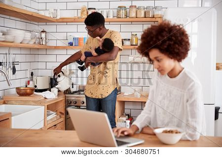 Multi-Tasking Father Holds Baby Son And Pours Drink As Mother Uses Laptop And Eats Breakfast
