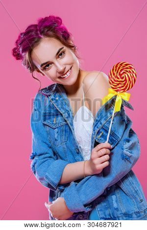 Pretty pink-haired girl with lollipop in denim jacket posing on a pink background. Fashion, beauty. Youth style of clothes.