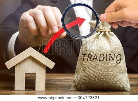 A Money Bag With The Word Trading, An Up Arrow And A Wooden House. The Concept Of Increasing The Dem