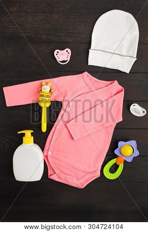 Pink Summer Bodysuit, Hat, Two Sylicone Pacifiers And Two Wooden And Plastic Rattles On Brown Wooden
