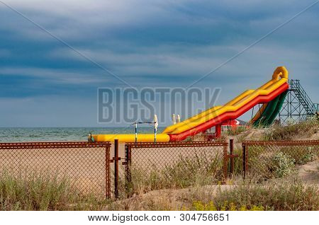 Colorful Inflatable Slide On Beach Sand Of Coastline. Seascape With Rusty Metal Fence And Aquapark E
