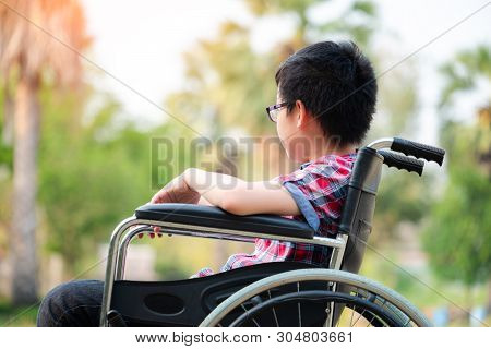 Alone Young Disabled Man On Wheelchair In The Park, Patient Is Relaxing In Garden Decorations Of The