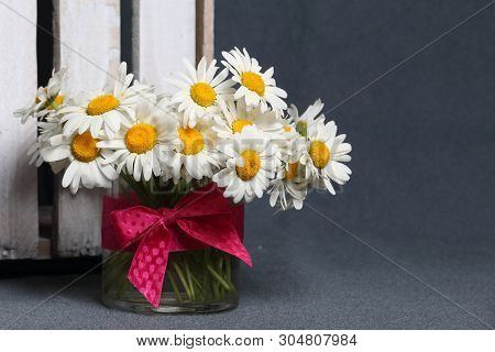 A Bouquet Of Field Daisies In A Glass Vase. Near The Wooden Box. Genus Of Perennial Flowering Plants