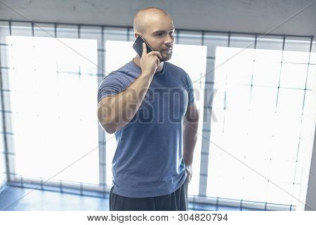 A Serious Sports Coach Speaks By Telephone With A Client In A Gym. Dressed In A Sports Uniform