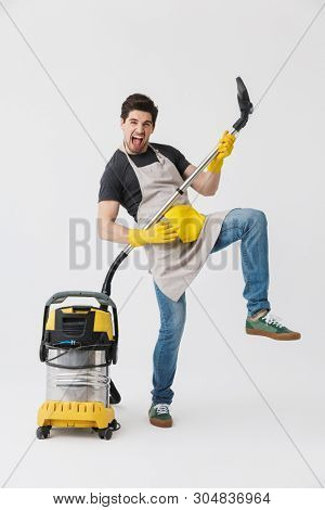 Full length of a handsome brunette houseman wearing apron standing isolated over white background, cleaning with vacuum cleaner