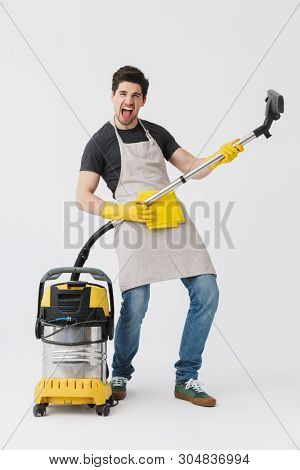 Full length of a handsome brunette houseman wearing apron standing isolated over white background, cleaning with vacuum cleaner