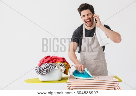 Handsome angry brunette houseman wearing apron standing isolated over white background, ironing clothes on a board, talking on mobile phone