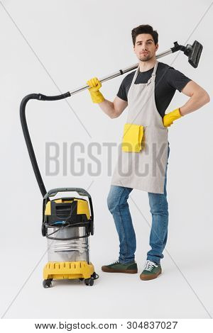 Full length of a handsome brunette houseman wearing apron standing isolated over white background, cleaning with vacuum cleaner